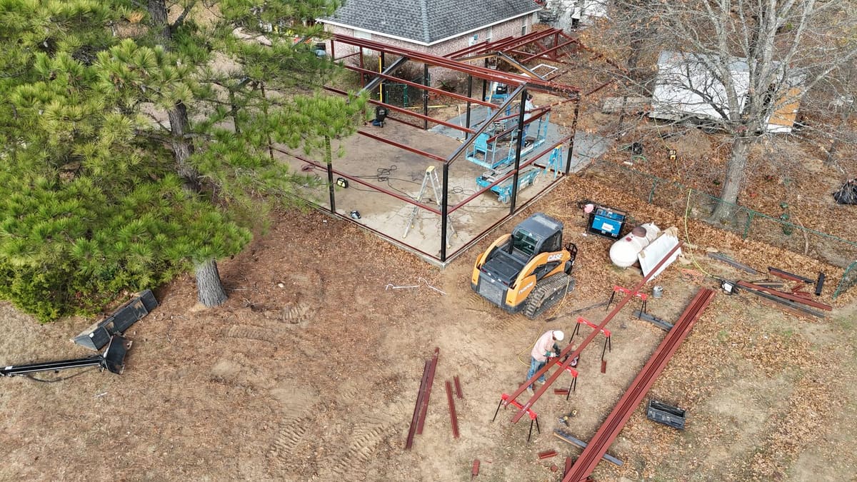 Aerial view of a steel frame structure being erected beside a home with a lift and crew — Red Moon Expert metal building contractor Texas
